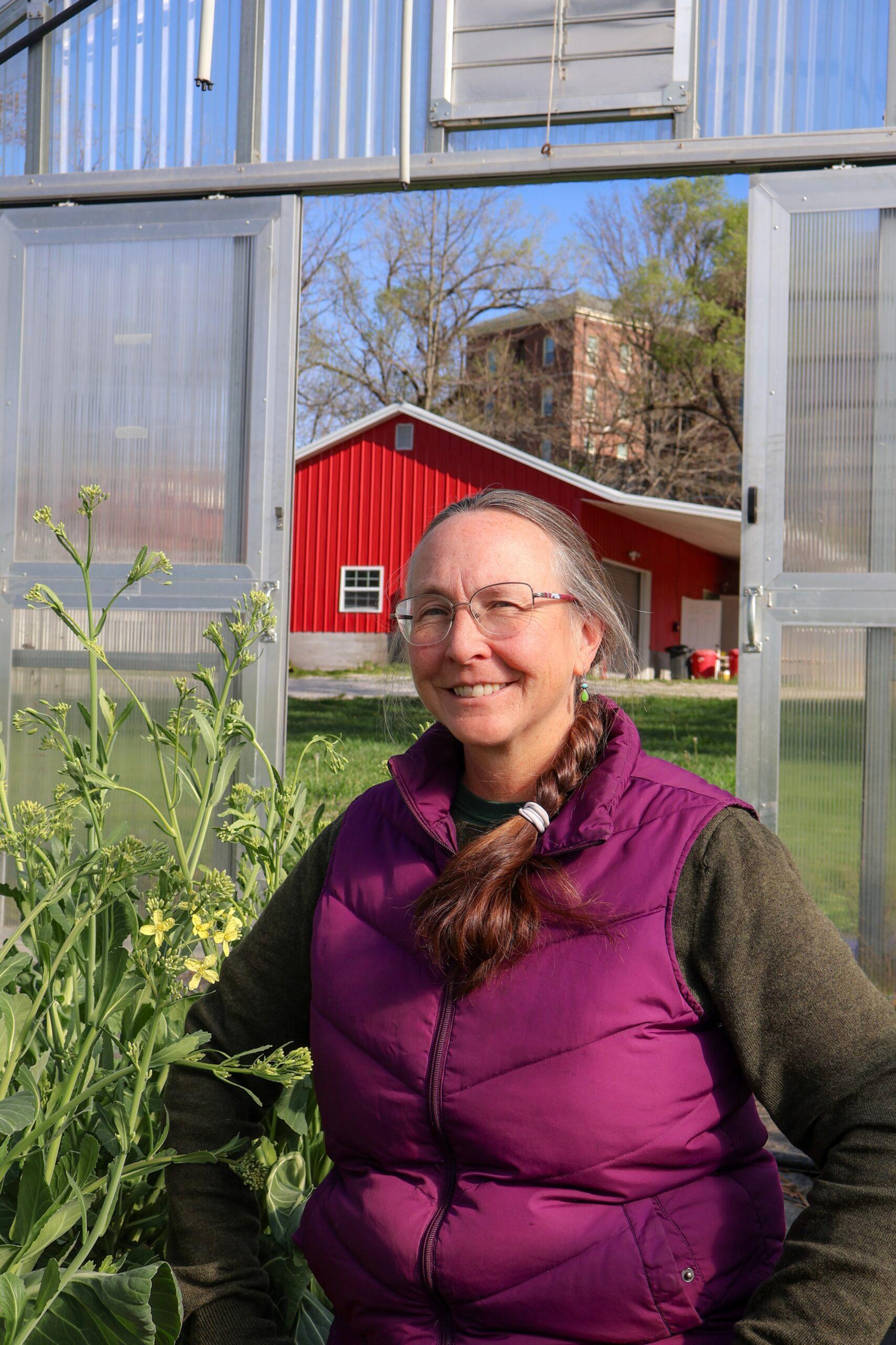 Janet sitting and smiling towards the camera, and near is cultivating plants, behind is a greenhouse door exit with nature and a red house. Janet is wearing a purple vest with a brown sweater.