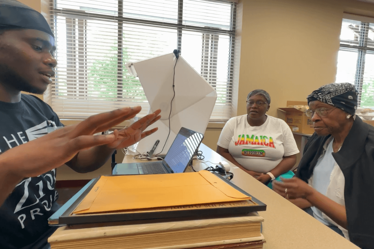 A male student, shown on the left wearing a black T-shirt, interviews two women, shown on the right, sitting across a table.