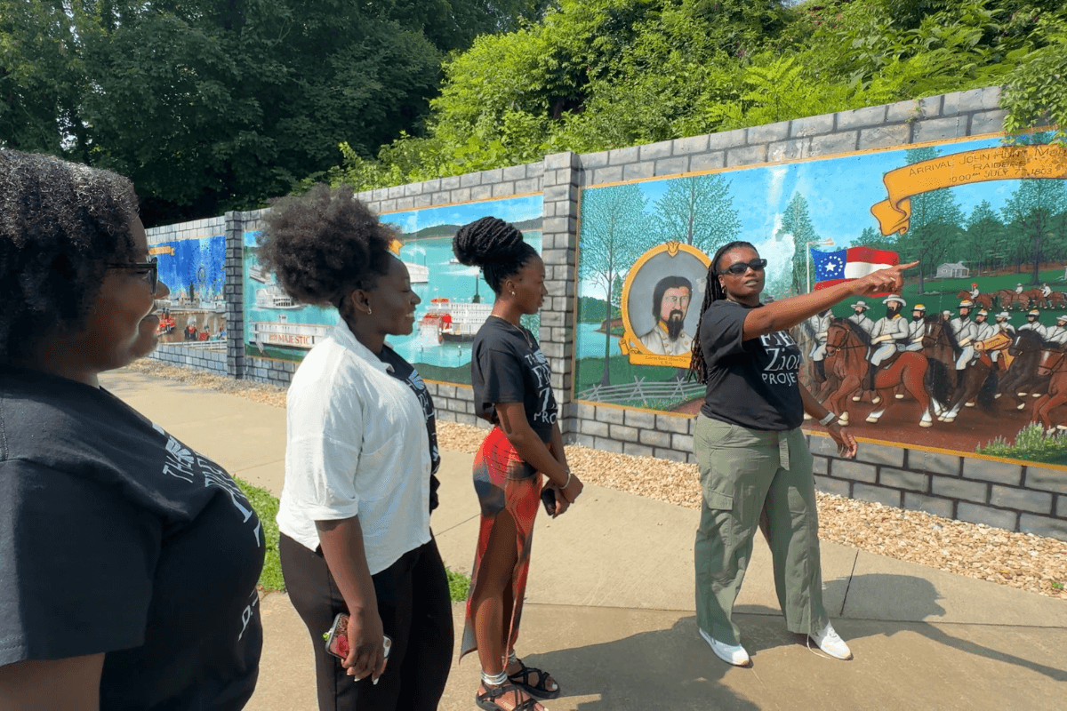 Four women stand outside in front of a painted mural looking off toward the right side of the screen.